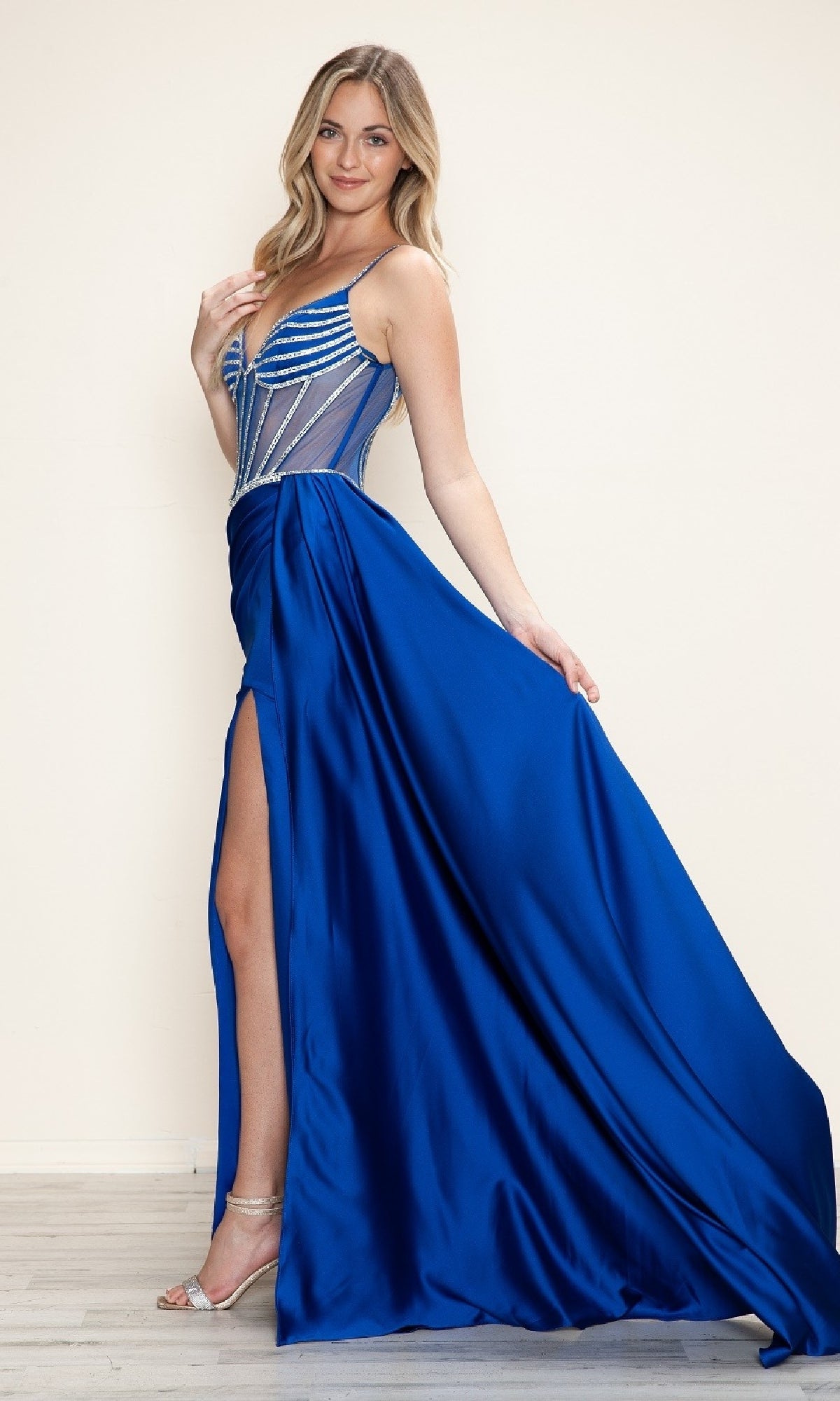 A woman poses on a wooden floor against a light background, wearing the Poly USA 9746 Beaded-Corset Long Prom Dress in Royal—a blue evening gown with a thigh-high slit and sheer, beaded corset bodice.